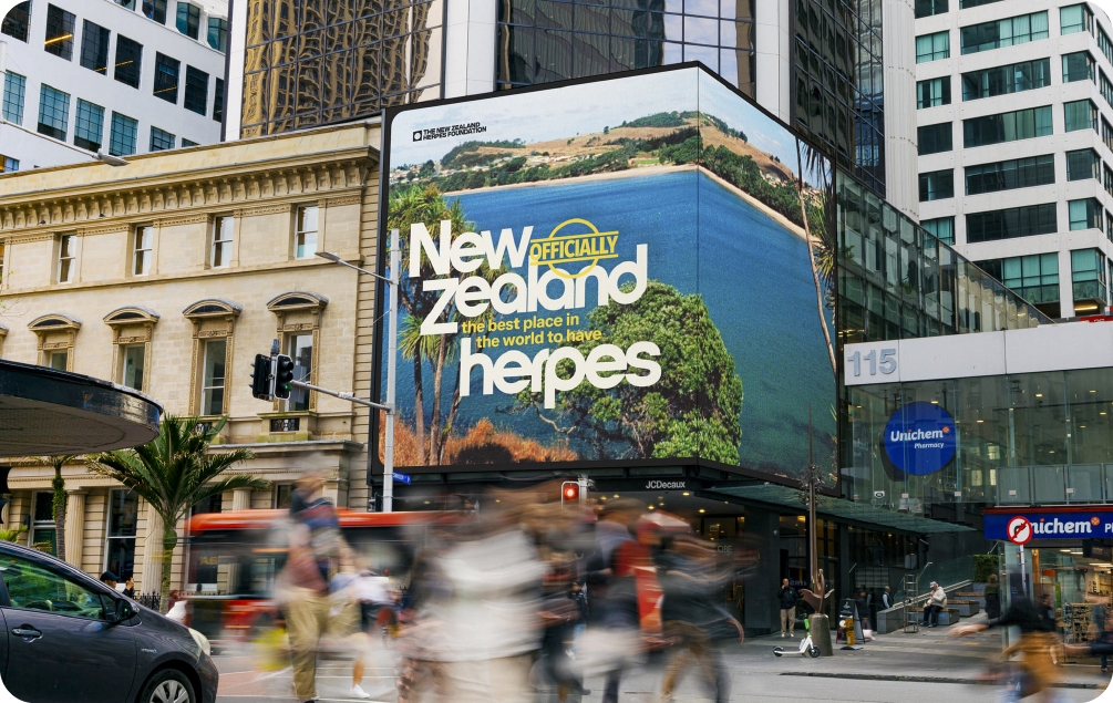 A large digital billboard on a city street corner in New Zealand displaying a scenic coastal image with the headline, "New Zealand officially the best place in the world to have herpes."